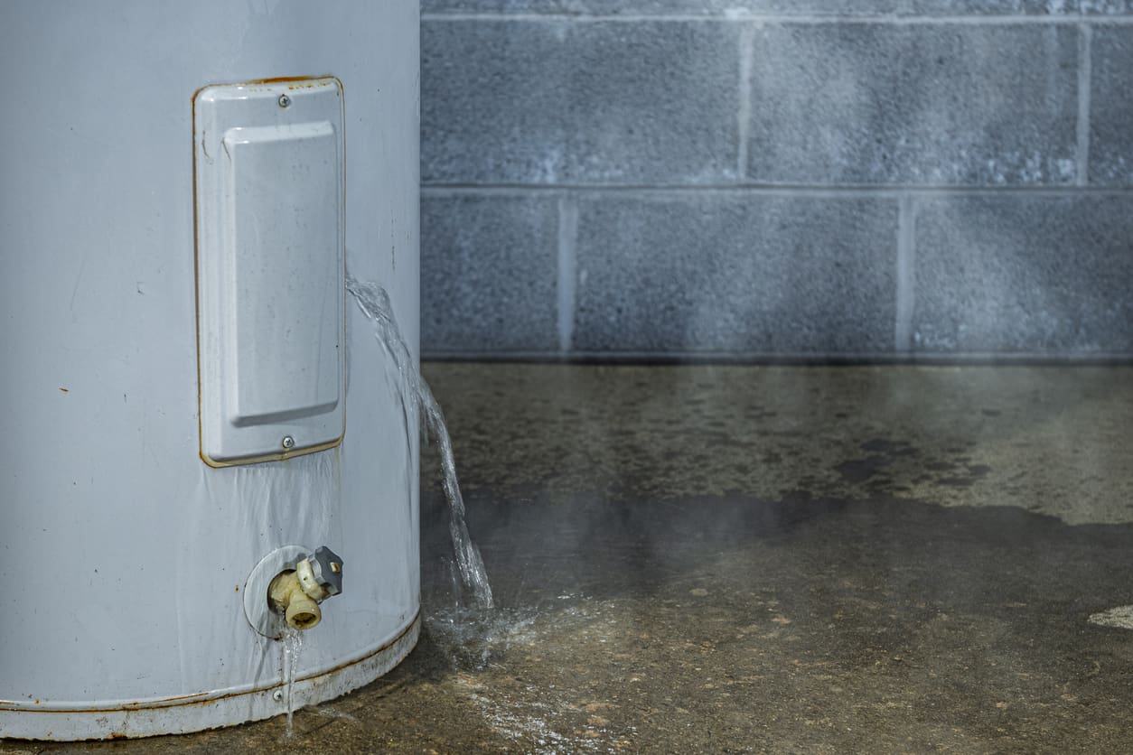 A close-up of water leaking out of an access panel of an electric water heater onto the concrete floor of a basement with a cinder block wall in the background.